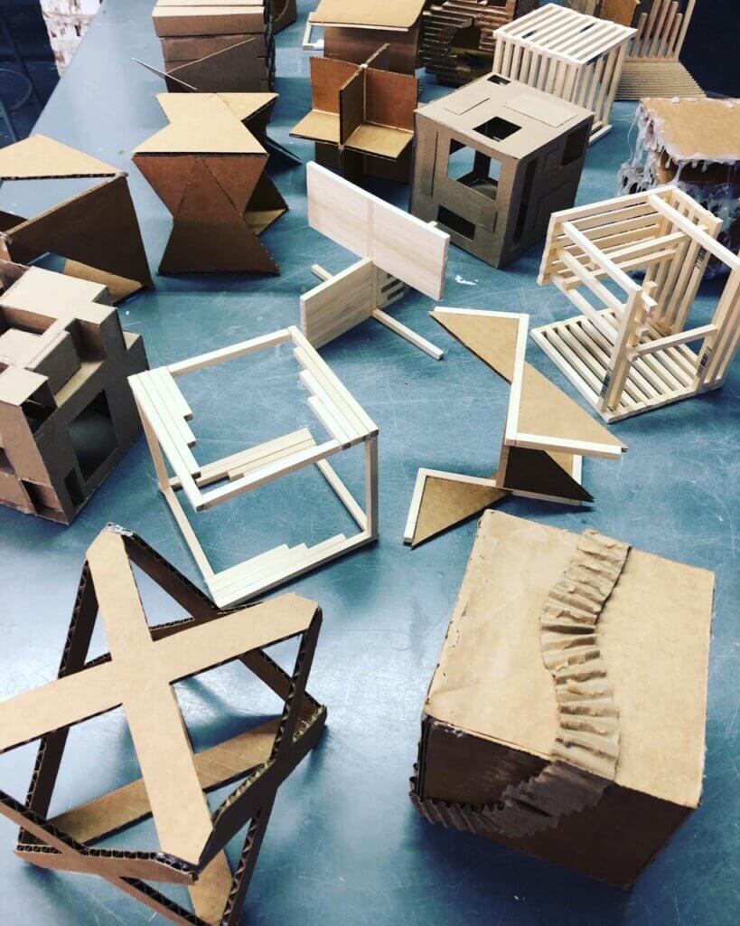 Collection of small abstract cardboard and wood structural models arranged across a blue tabletop.