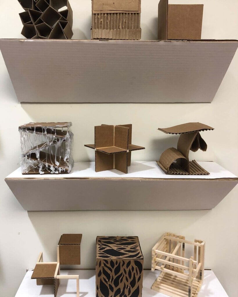 Display of small cardboard and wood sculptural models arranged on white wall-mounted shelves.