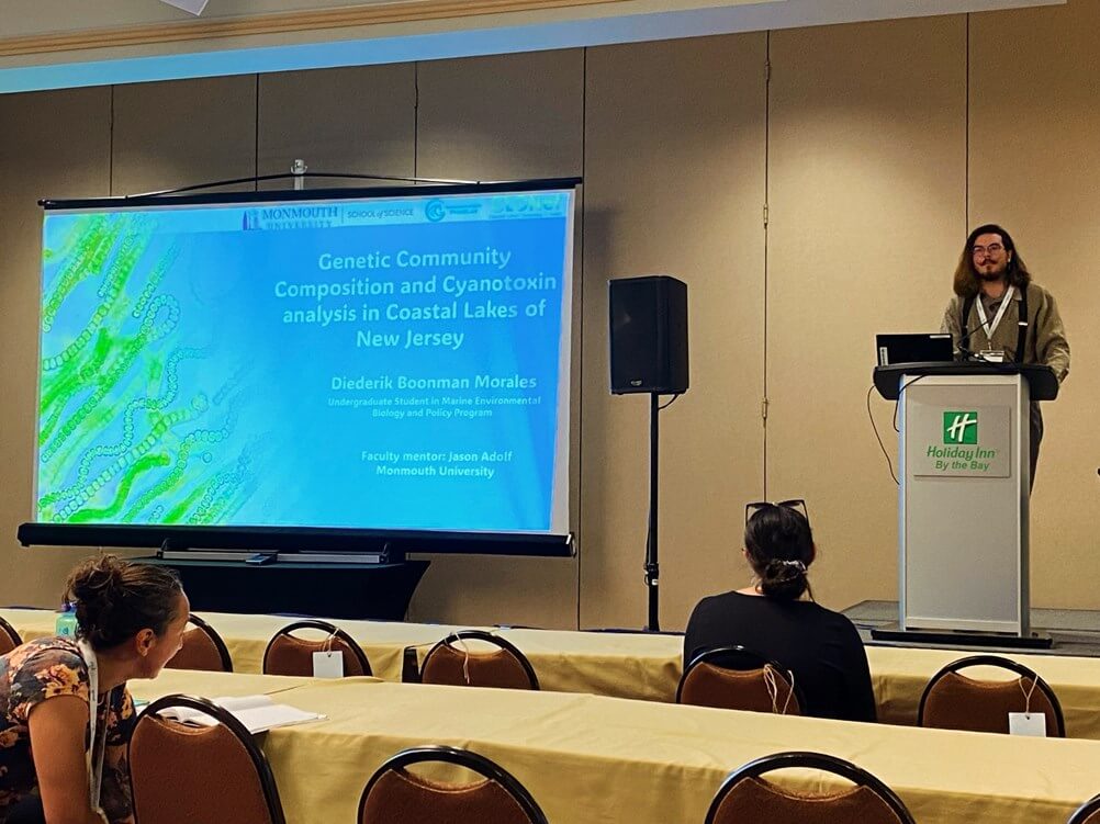 Man at podium, with his presentation projected on a screen. The title reads "Genetic Community Composition and Cyanotoxin Analysis in Coastal Lakes of New Jersey