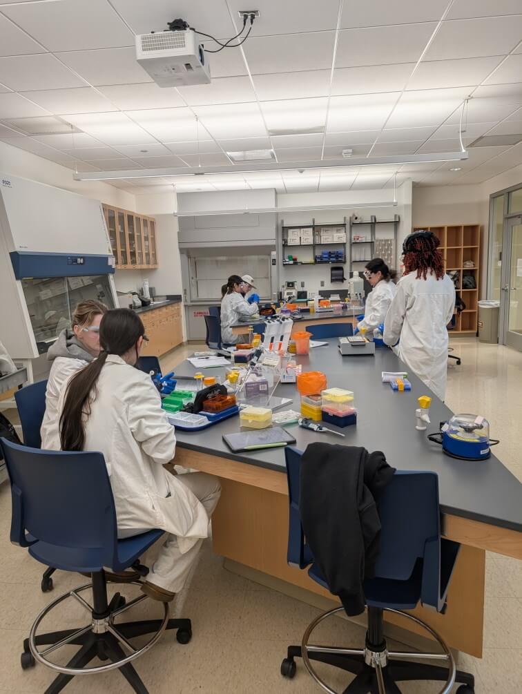 Students talking amongst themselves, all wearing white coats, in side a lab