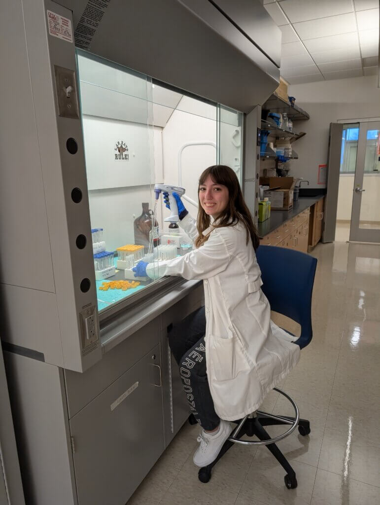 Woman in white coatworking with equipment housed inside a cabinet with glass panels