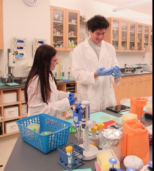 A man and a woman in white coats putting on rubber globes near table filled with scientific equipment