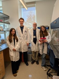 Three students and a professor in a lab, all wearing lab coats. 