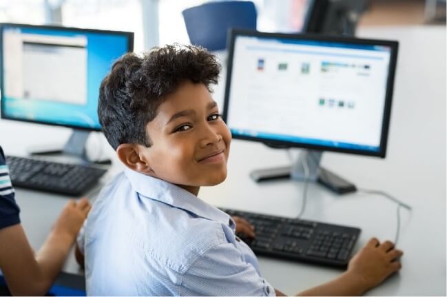 A child is smiling at the camera while working with a computer.