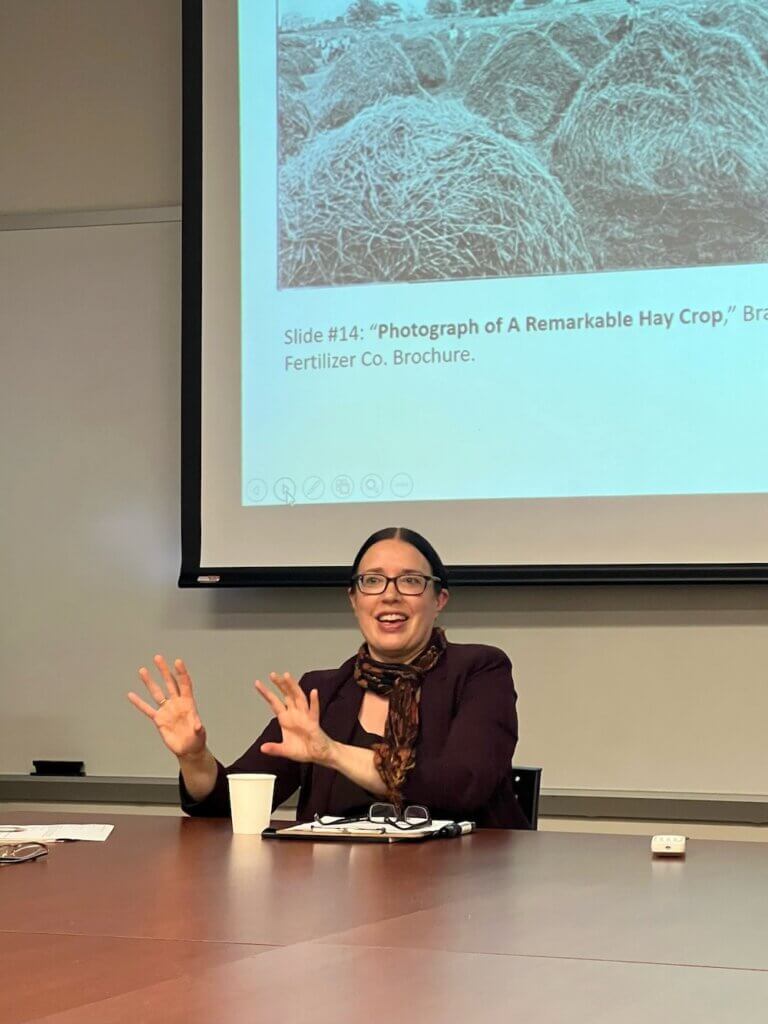 Woman speaking from a table, presentation projected on a canvas in the background