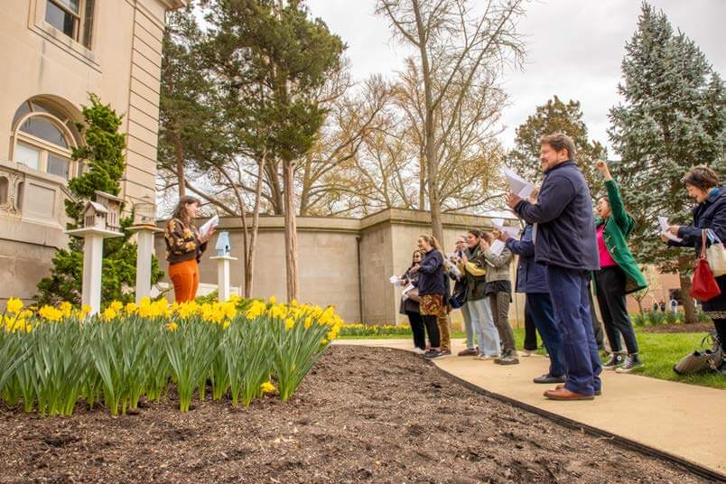 Woman reading from a book in a garden, with audience listening