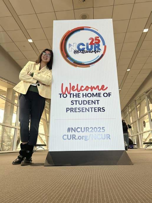 Woman leaning on freestanding sign that reads "25 NCUR: Welcome to the Home of Student Presenters"