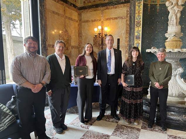 Six people posing for photo in ornate room, a fountain behind them