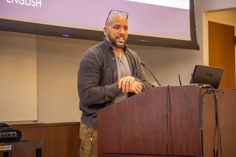 Man stands at podium with one hand over the other, glasses resting on forehead