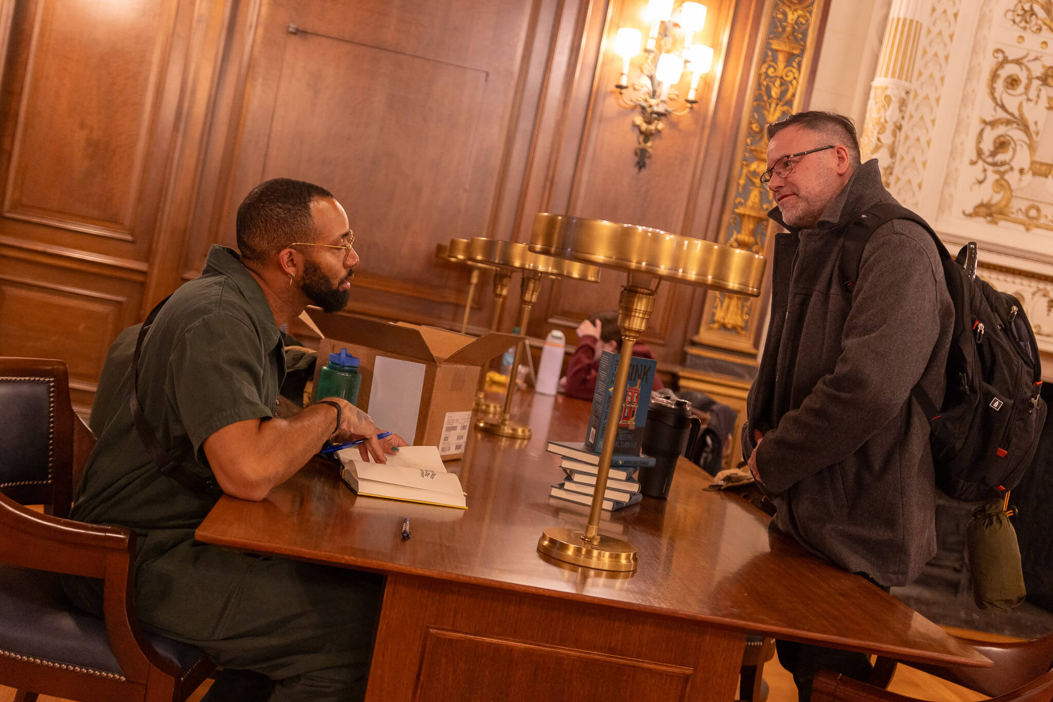 Man sitting down with book open on table, talking to another man across the table