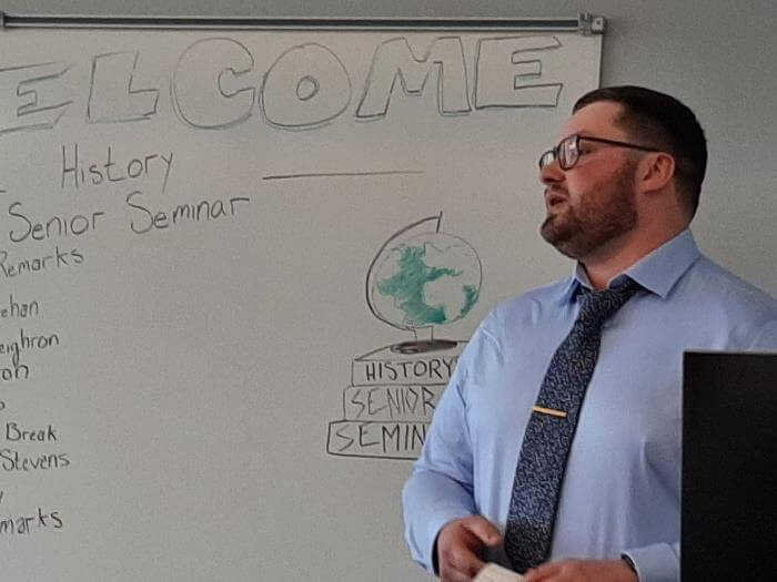 Man in blue shirt and tied speaking in front of a whiteboard that reads "Welcome. History Senior Seminar"