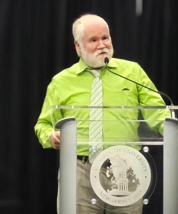 Man in green shirt and tie speaking at podium with Monmouth University seal