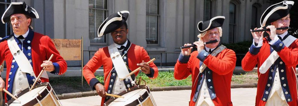 Man in colonial marching band outfits, two men on drums, two on flutes.