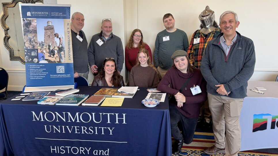 Group of students and teachers at Monmouth University table during history conference, one wearing a steel helment