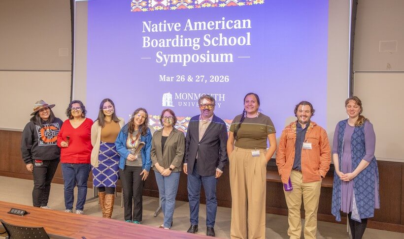 Row of men and women standing in front of a screen reading "Native American Boardin School Symposium, March 26 and 27, 2026, Monmouth University."