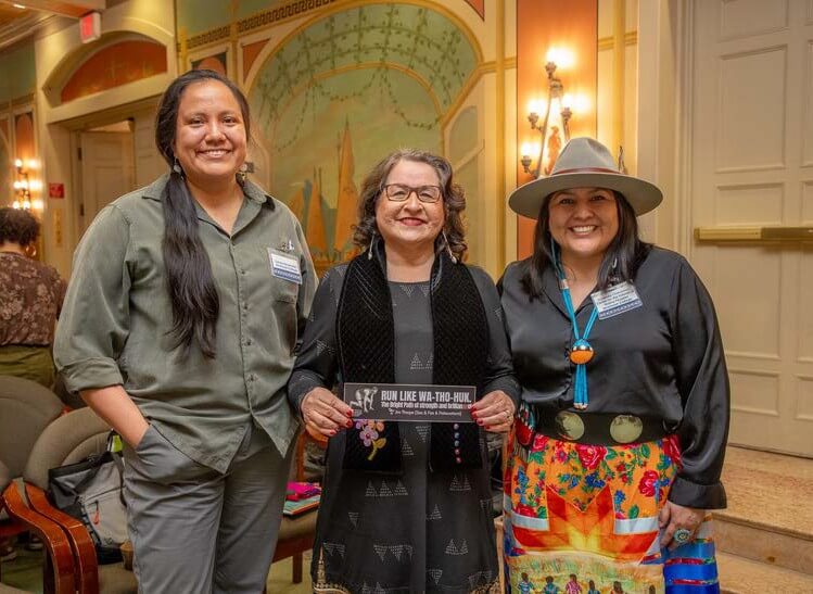 Three women smiling for camera, one holding a sign reading "Run like Wa-Tho-Huk"