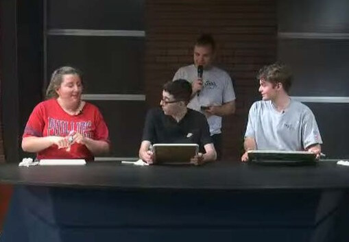 Three people at table speaking, while a man with a microphone stands behind them.