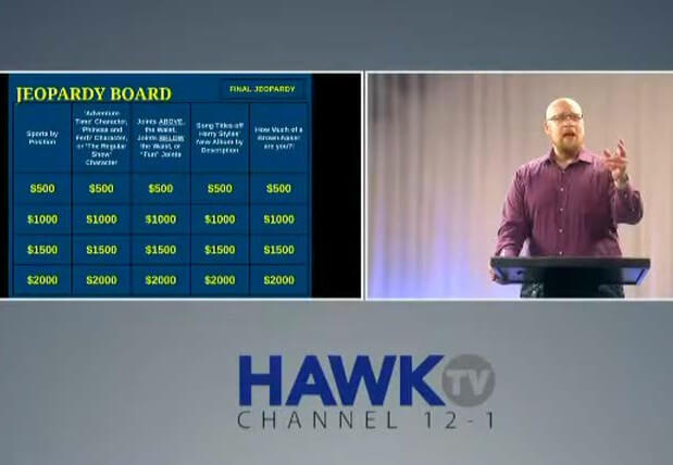 Screenshot of tv show. Man at podium, a jeopardy board presented next to him. The Hawk TV logo appears at the bottom