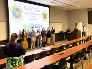 Students in front of screen welcoming them into their honors socieity. Their hands are raised for their induction.