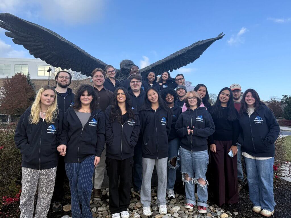 Young adults in coordinated Debate Team hoodies standing near a hawk statue