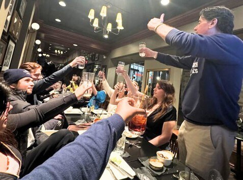 Man standing up at restaurant tabl giving a thumbs up while toasting his drink, the other folks at the table also toasting.