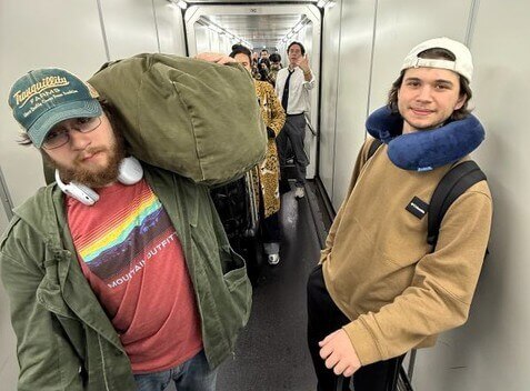 Two students walking through narrow plane terminal hallway, one with headphones around neck, the other with neck pillow. Res of group is behind them.