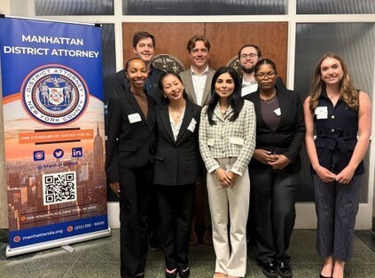 Group of adults in suits posing for photo by banner for Manhattan District Attorney