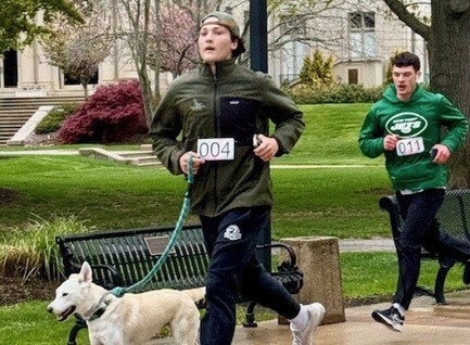 Two people running on a campus, one with a dog on a leash.