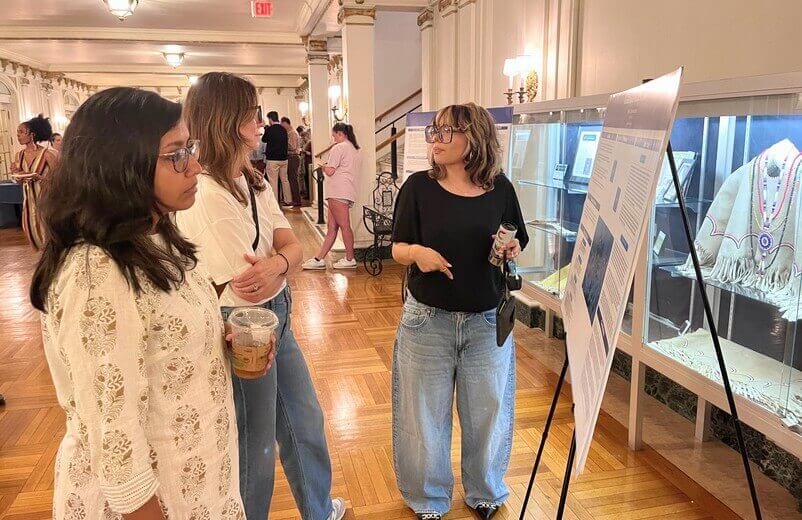 Student near poster presentation speaking with attendees in a hall
