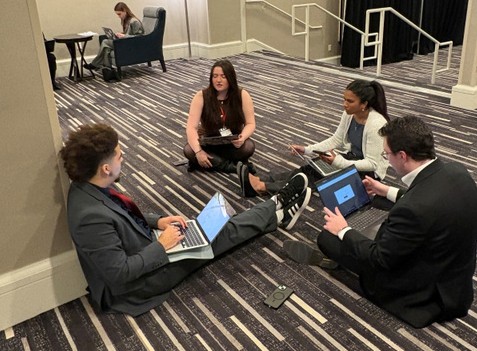Four conference goers sitting on a carpeted floor, some on laptops, speaking with each other