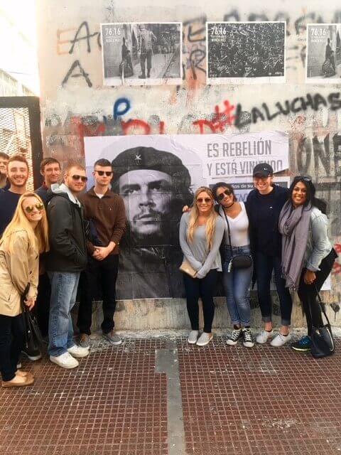 Group of people standing near poster of Che Guevar on wall with graffiti and posters