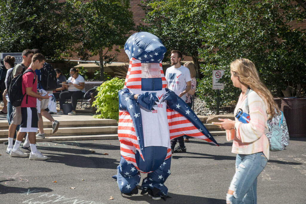Person in red white and blue dinosaur costume talking to students during a Rock the Vote event.