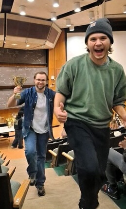 Two debaters smiling and walking up lecture hall steps, one holding a trophy and the other giving a thumbs up
