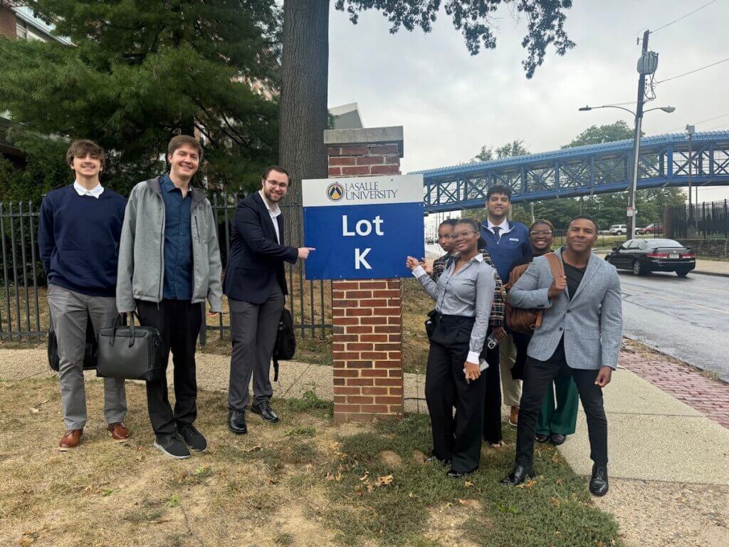Students on a lawn wearing suits and business attire, pointing at a sign reading Lot K.