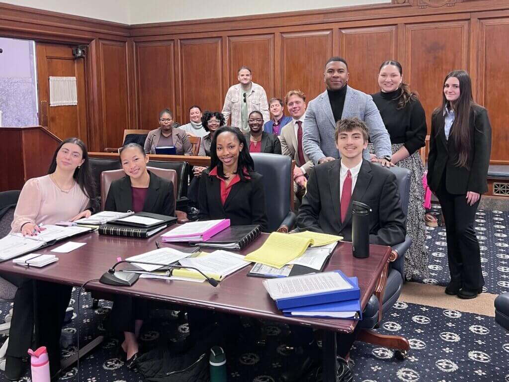 Students in a court room like setting, some sitting at a lawyer's table, others in the back