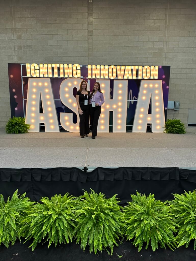 Two women in front of a large sign that says Igniting Innovation ASHA