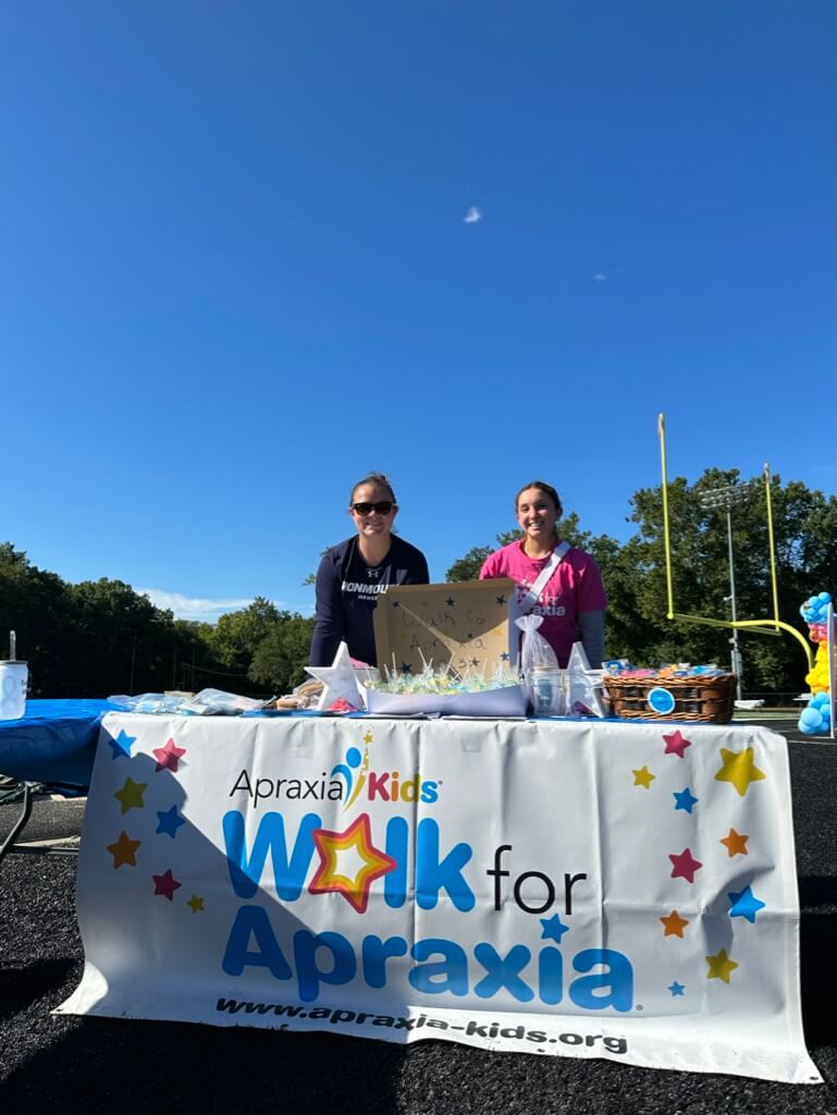 Two women on a football field behind a table with a sign that says Apraxia Kids, Walk for Aprakia