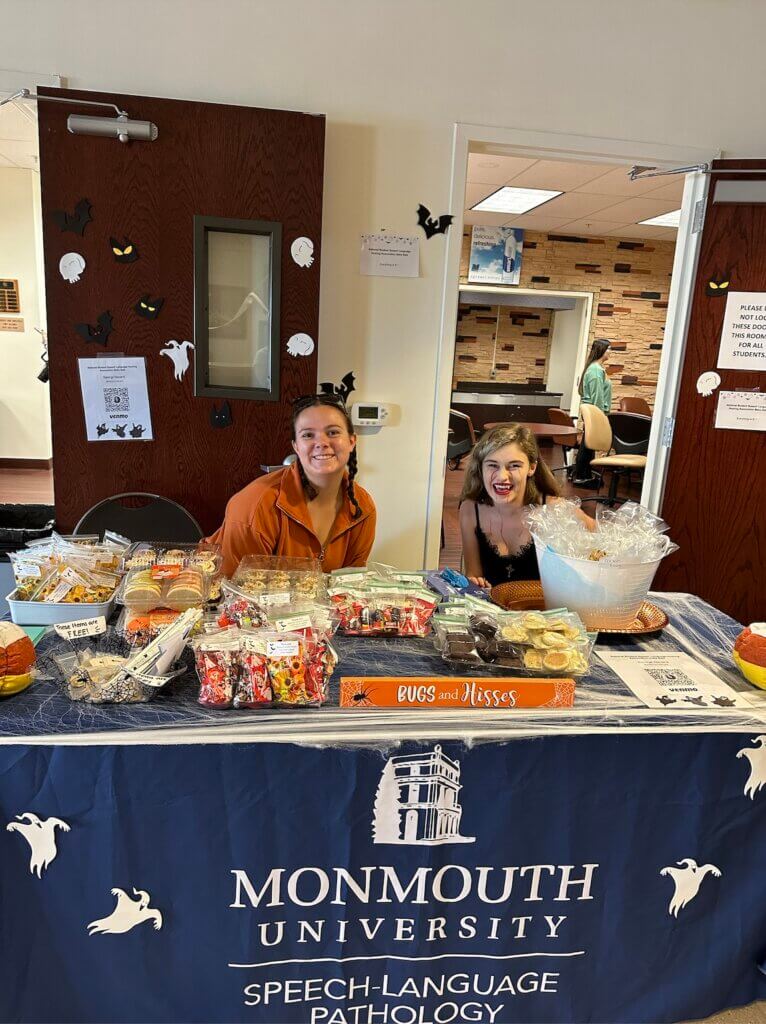 Two women, one is in Halloween makeup, are sitting behind a table with candy on it. A banner with the Monmouth Logo and text 'Speech-Language Pathology' is draped over the table. 