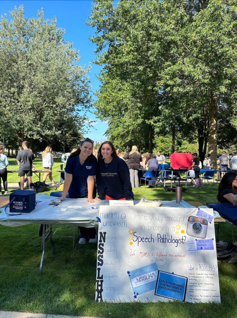 Two women are standing at an informational table promoting NSSLHA while outside on a sunny day with other tables promoting other clubs