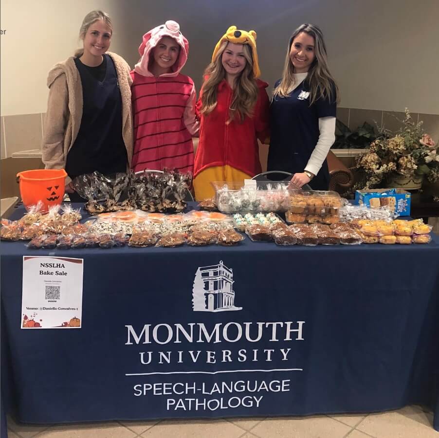 Four women standing behind a table for a bake sale. Two of the women are wearing Winnie-the-Pooh themed onesies.
