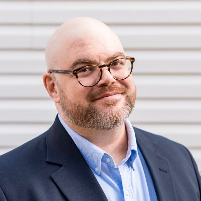 A headshot of Ryan Eckert who is wearing a light blue shirt and dark jacket, he has glasses on and a short beard.