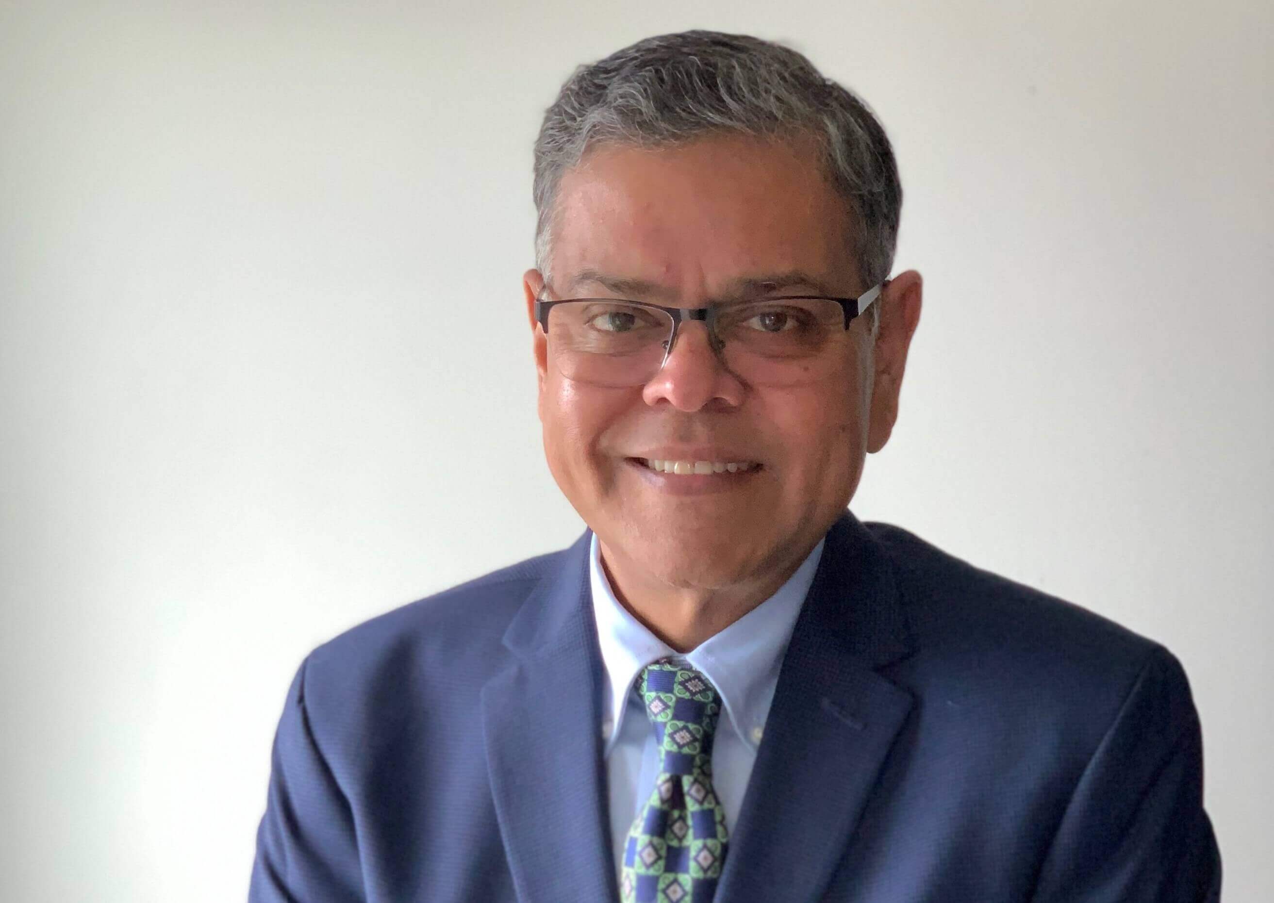 A head shot photo of Raj Devasagayam, he is infront of a white background while wearing a blue suit and tie.