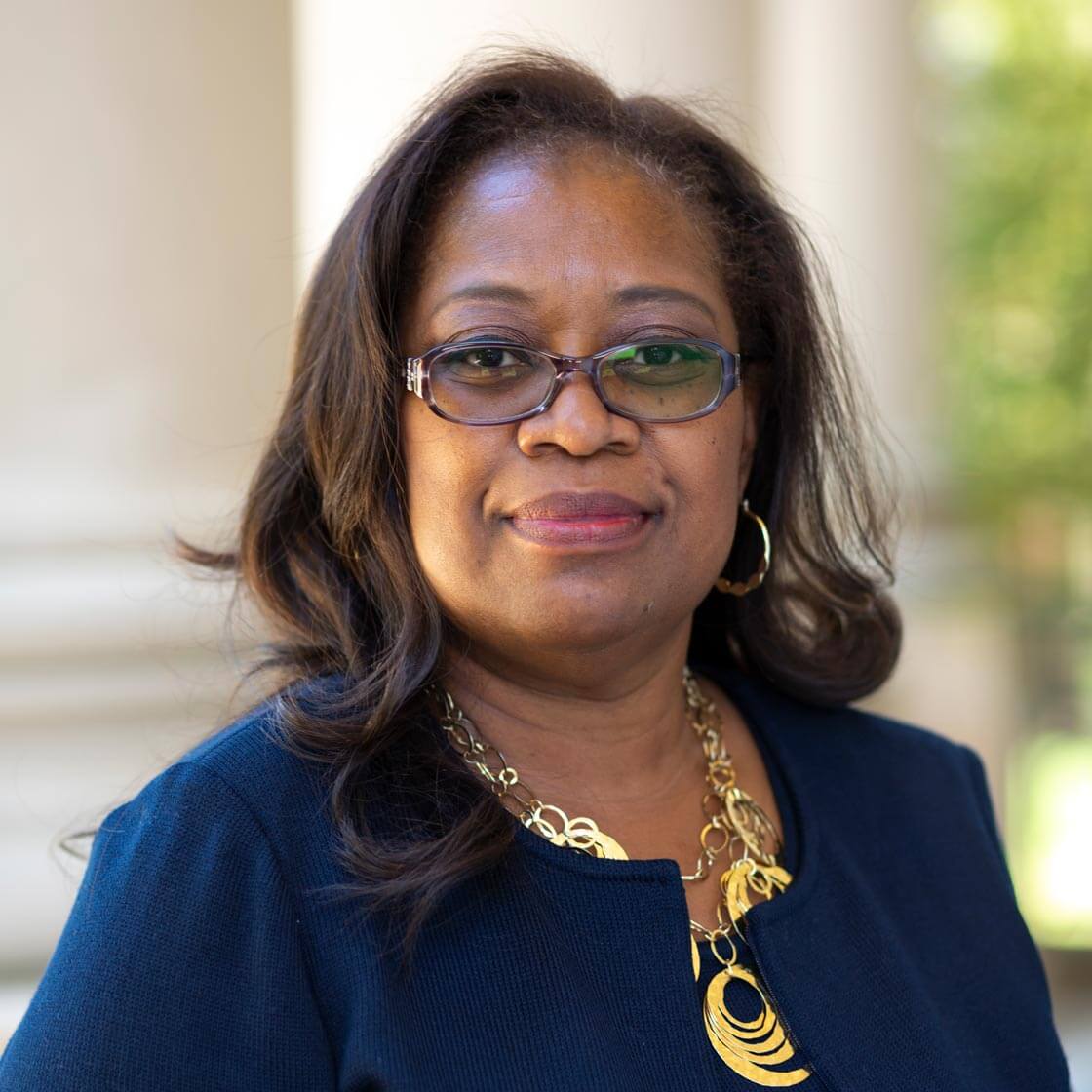 A head shot of Pamela Scott-Johnson, who is wearing glasses, has sholder length hair, and is also wearing a blue jacker with a gold necklace made up of various sized circles