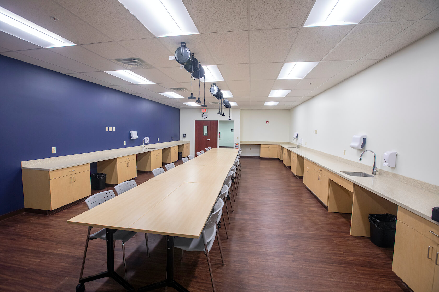 A room with a large table, chairs arranged underneath. There's a series of sinks arranged on two countertops.