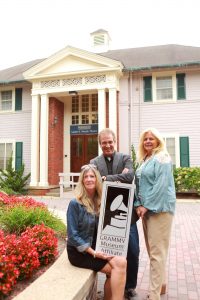 Three people in front of the Wood Theater holding a Sign that has the GRAMMY Museum logo on it