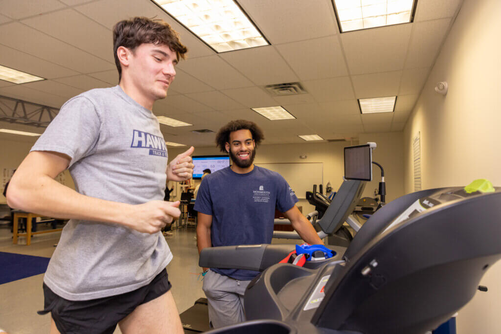 A man in a blue shirt watches another man run on a treadmill. Other exercise equipment appears in the background