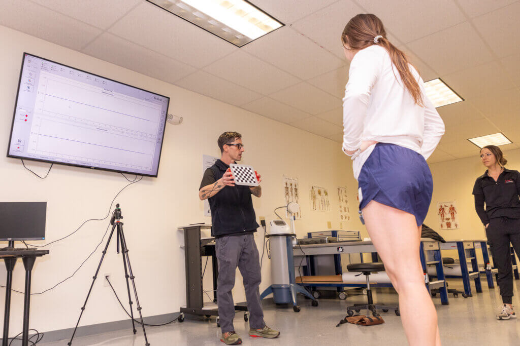 A man shows a display with a checkberboard pattern to two women. Lab equipment and human anatomy posters appear in the background