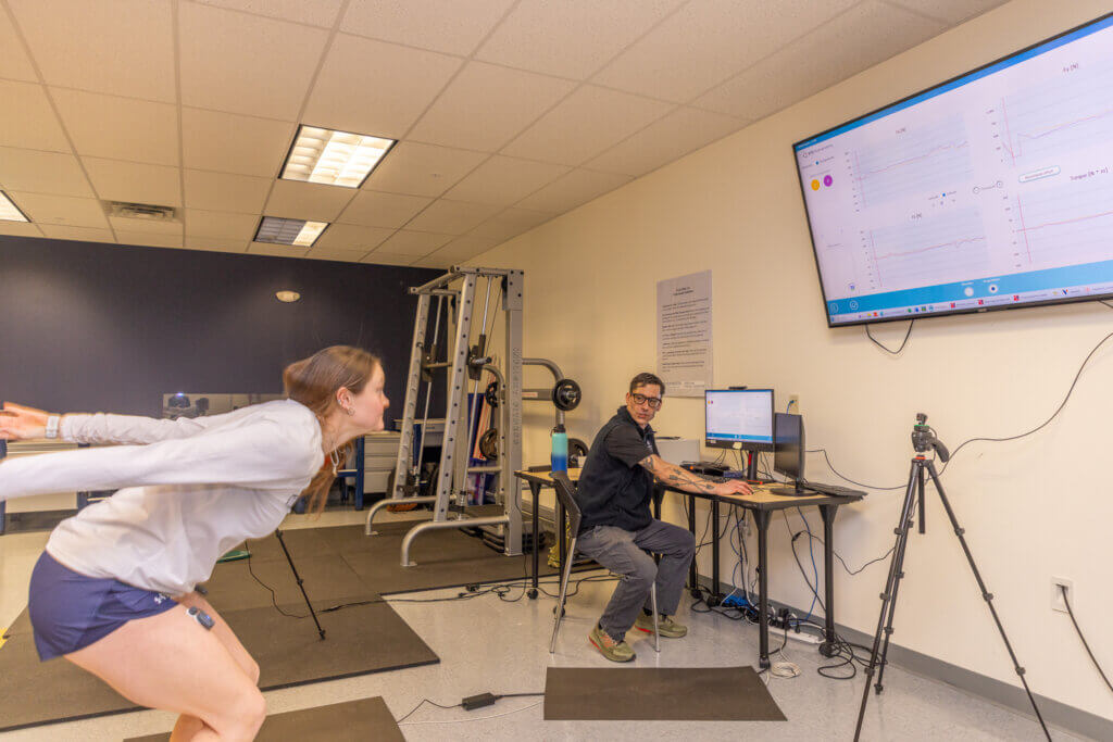 A man on a computer looks on as a woman performs exercises. The woman is wearing equipment for analytical purposes.