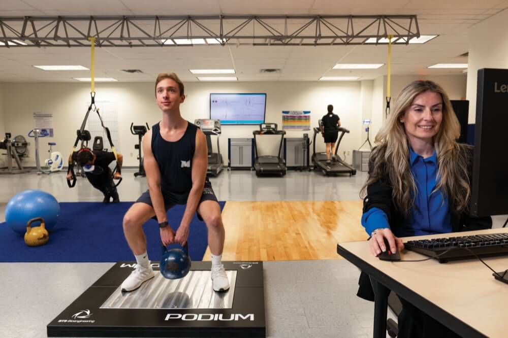 Man crouching, holding weight while standing on a scale, while a woman uses a computer nearby.