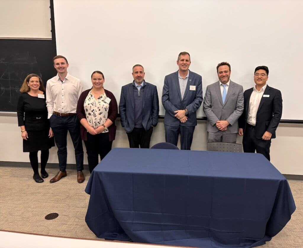 Two women and five men in suits, standing in front of a classroom blackboard, smiling for the camera